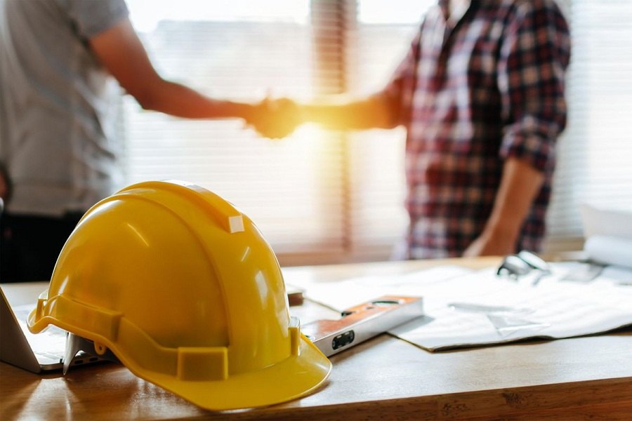 Chủ nhà bắt tay nhà thầu đội mũ bảo hộ bên cạnh bản vẽ thiết kế